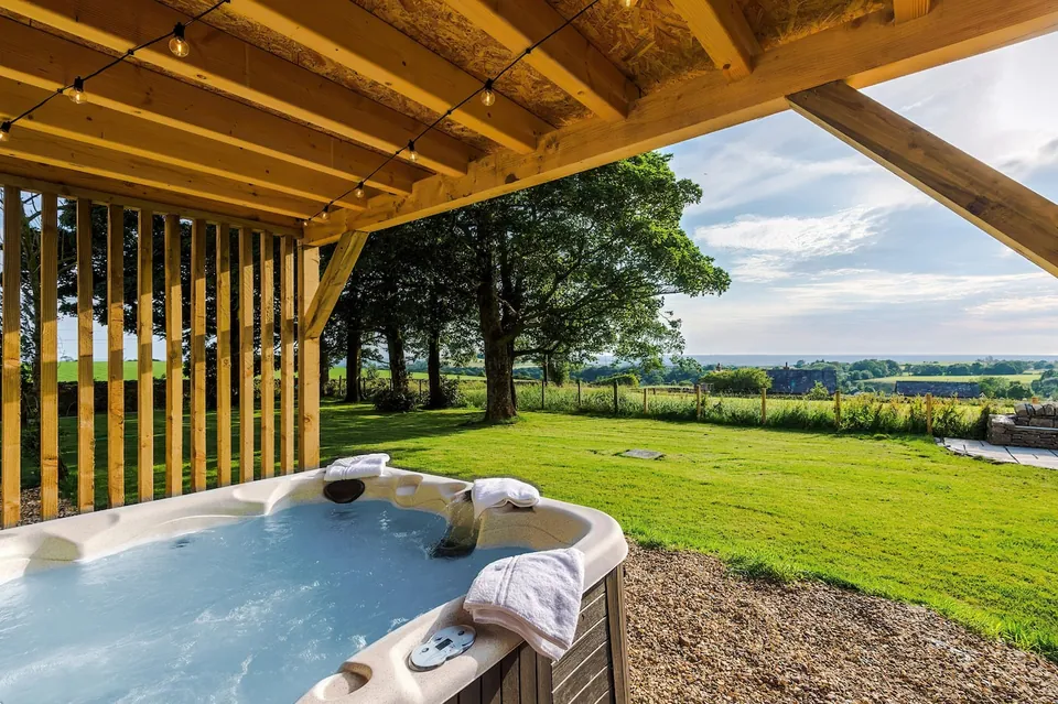 A hot tub with towels on the rim, under a wooden shelter strung with lights, facing a lawn and trees.
