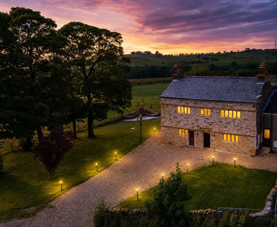 The stone house at dusk, with lights in the windows and along the drive, under a pink and purple sky.